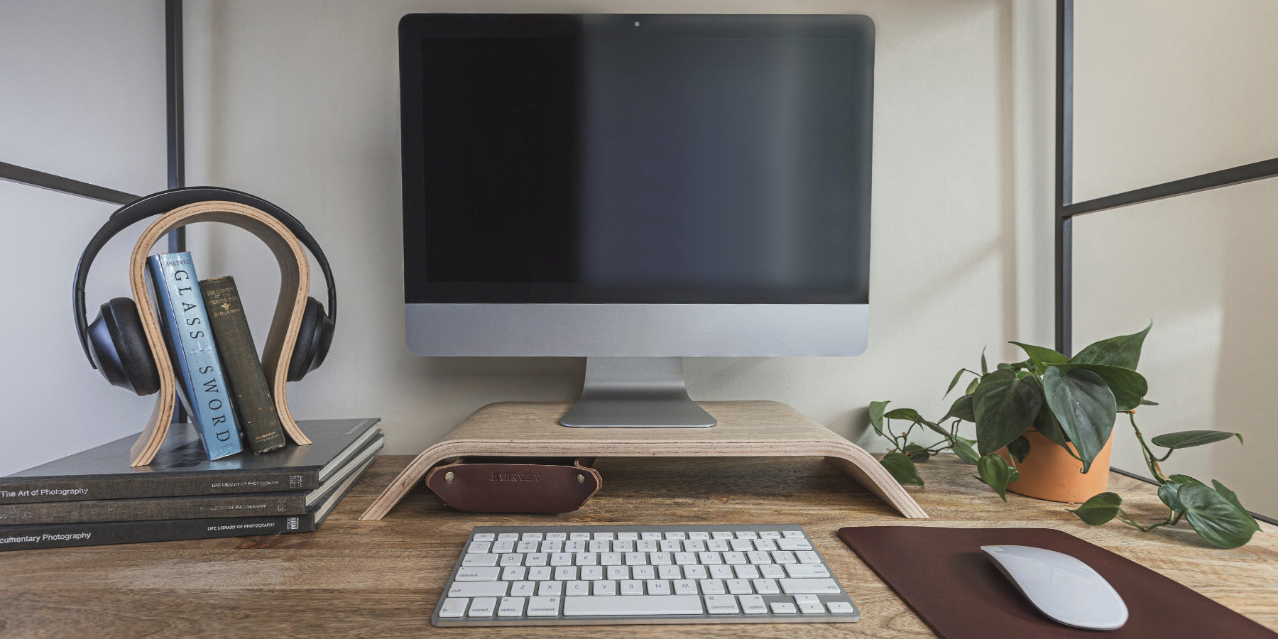 A minimalist home office setup featuring a large computer monitor on a wooden stand, with a wireless keyboard and mouse on a brown desk mat in front. To the left, a headphone stand holds a pair of black headphones, with several books stacked underneath. On the right side, there is a potted plant adding a touch of greenery to the workspace.