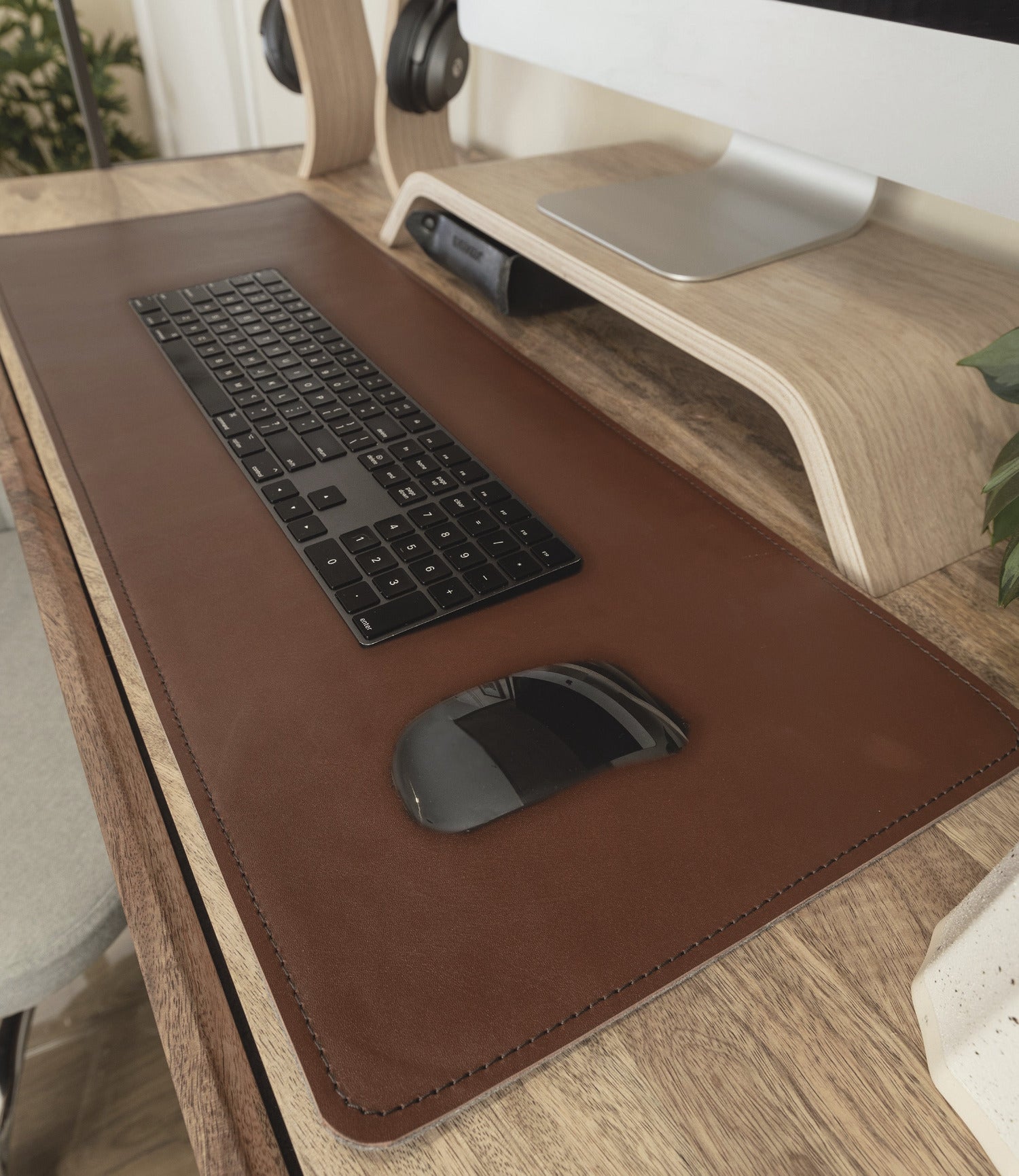 A close-up view of a sleek, minimalist desk setup with a brown leather desk mat. On the mat, there is a black wireless keyboard and a matching black wireless mouse. In the background, there is a wooden monitor stand holding a computer monitor, with a pair of headphones on a wooden headphone stand to the left. A small plant is partially visible on the right side of the desk.