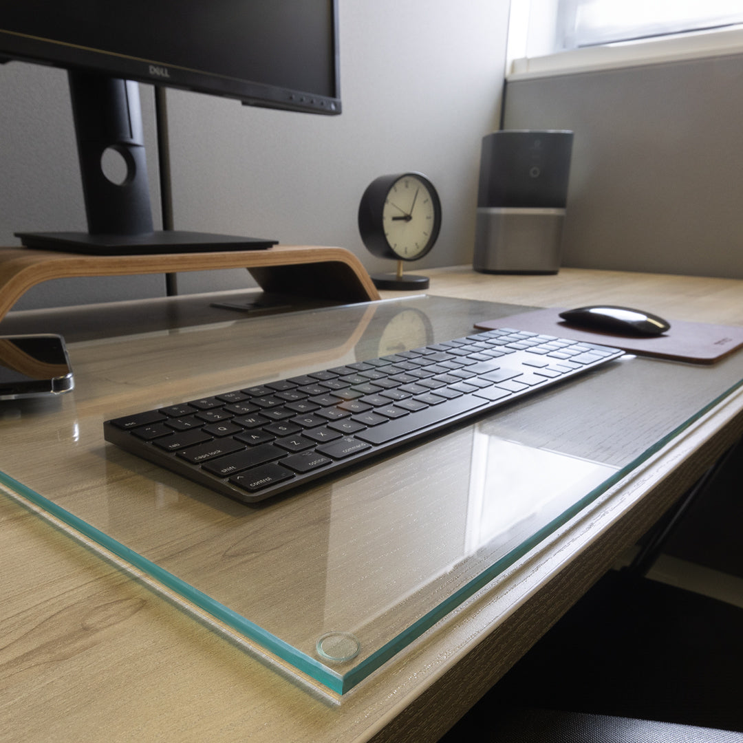 #glass clarity_chiaro (standard clear) alt="A clean and organized desk setup with a monitor on a wooden stand, a wireless keyboard on a "Chiaro" glass desk blotter, a mouse on a leather mouse pad, a smartphone, a round analog clock, and a cylindrical speaker. The background features a light-colored wall and a window letting in natural light."