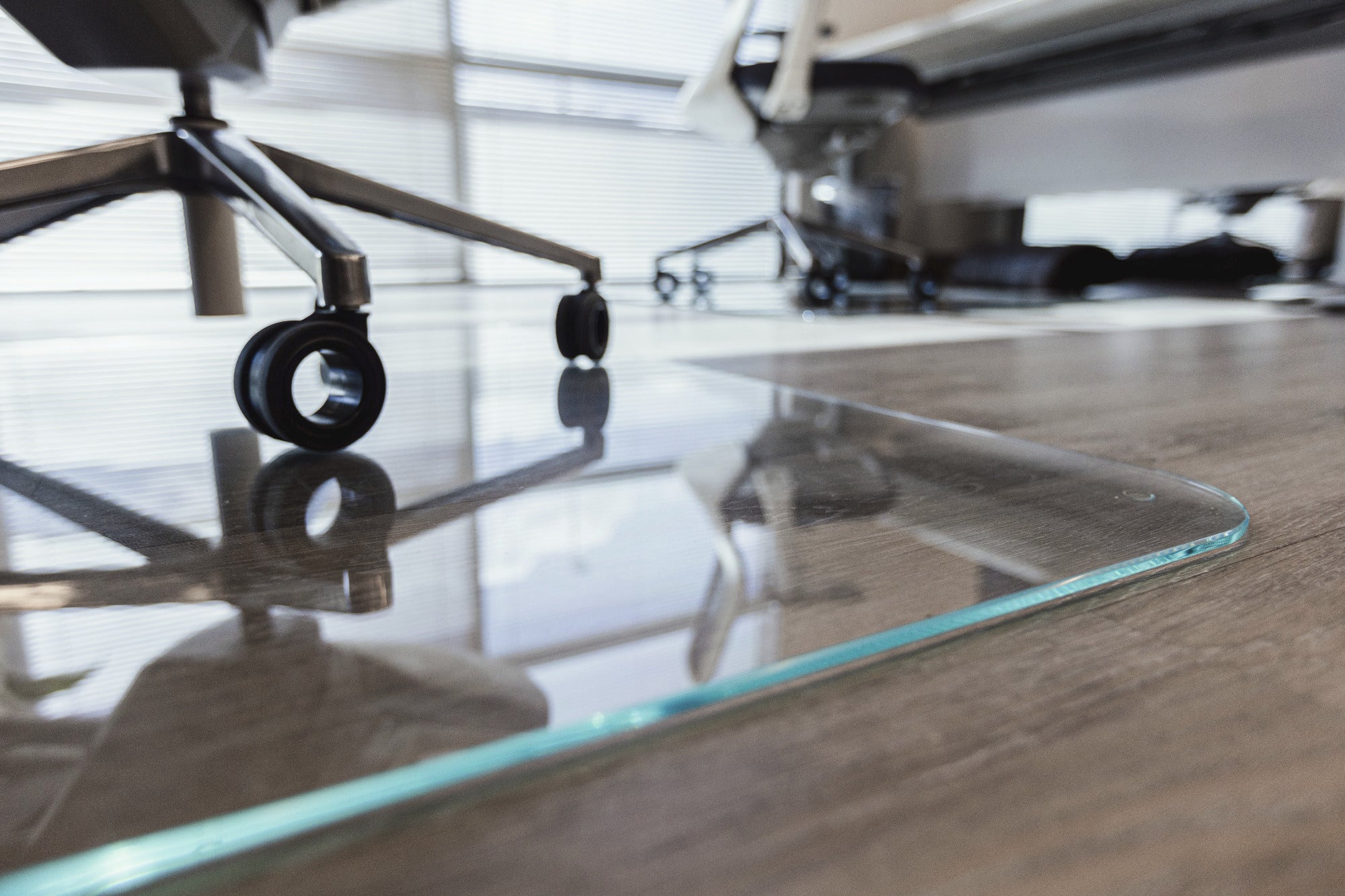 A close-up view of a clear glass desk mat on a wooden desk, with the wheels of an office chair resting on it. In the background, there are more office chairs and a window with blinds, suggesting an office environment.