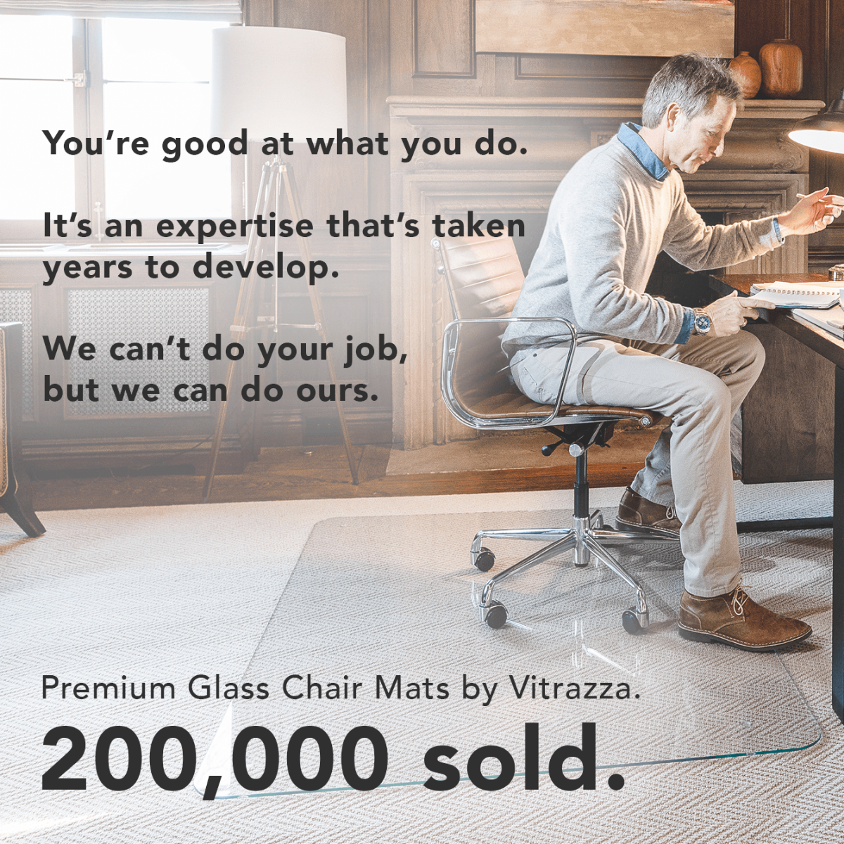 An advertisement for Vitrazza's Premium Glass Chair Mats, featuring a person working at a desk with the text: "You're good at what you do. It's an expertise that's taken years to develop. We can't do your job, but we can do ours. Premium Glass Chair Mats by Vitrazza. 200,000 sold." The setting is a well-lit, cozy office with a glass chair mat under a modern office chair.